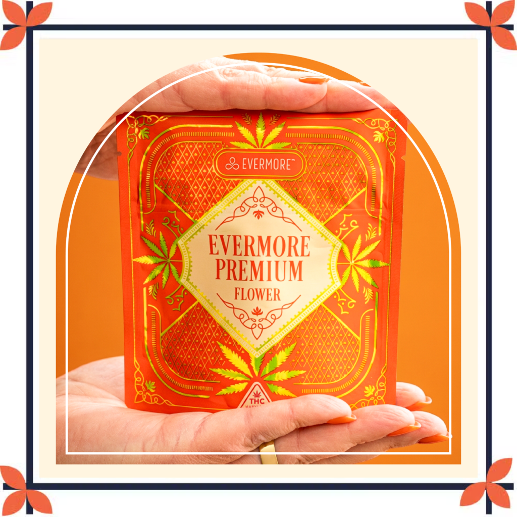 A bright orange Evermore Premium Flower cannabis package is held between two hands against a warm orange background. The package features intricate gold decorative details, cannabis leaf illustrations, and a cream-colored center label with the text "Evermore Premium Flower." The image is framed with a cream and navy border accented by orange floral corner motifs, creating a vintage-inspired design.