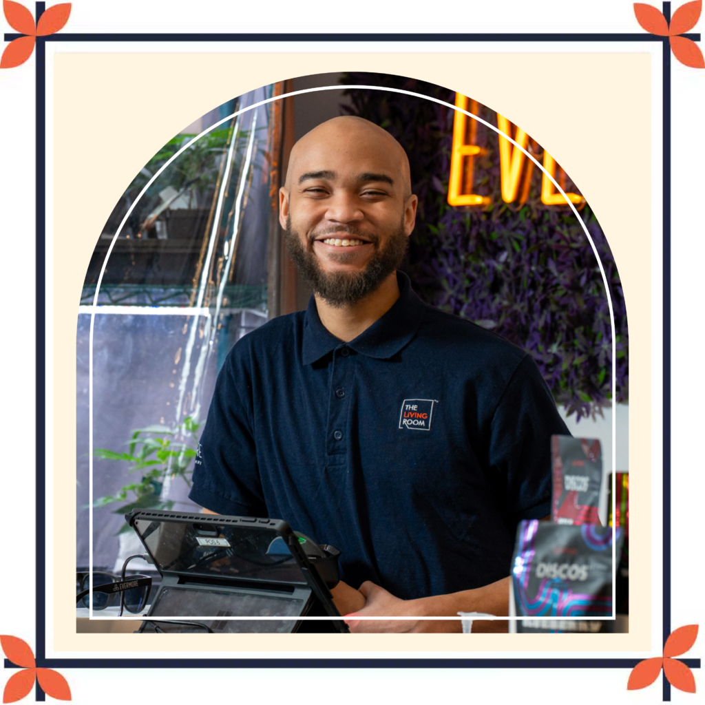 A smiling dispensary team member stands behind the checkout counter at The Living Room Dispensary, wearing a navy polo shirt with The Living Room logo. A point-of-sale tablet sits on the counter in front of them, while plants and product displays are visible nearby. The warm, welcoming interior features a glowing neon sign and decorative greenery in the background, creating a friendly and inviting atmosphere.